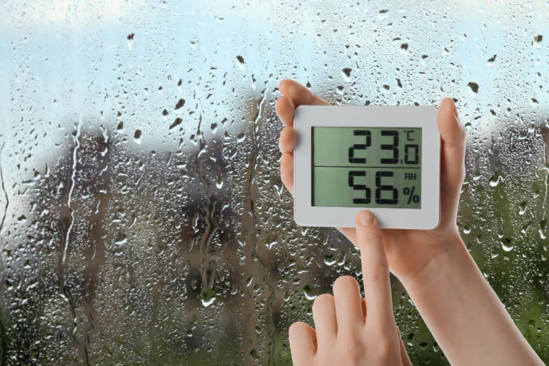 Digitální vlhkoměr ukazuje relativní vlhkost vzduchu před oknem s kapkami vody.