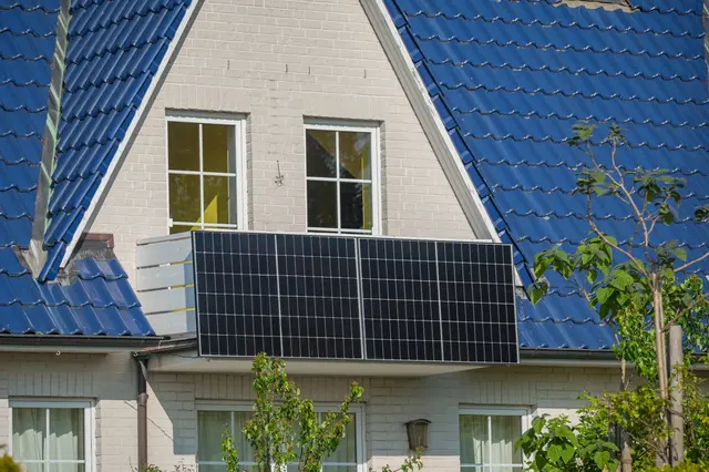 Fotografie fotovoltaických panelů na balkóně rodinného domu s modrou střechou.