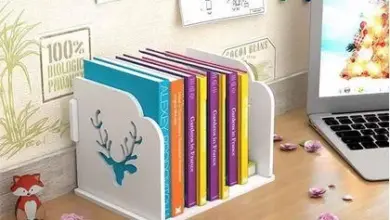 Bílá dekorativní knihovna s barevnými knihami na dřevěném stole vedle notebooku.