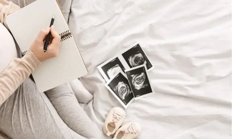 Těhotná žena píše do sešitu vedle ultrazvukových snímků a dětských botiček.